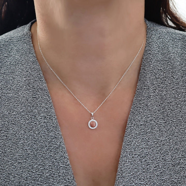 A close-up of a person wearing a delicate silver chain necklace with a small circular pendant featuring a pink rhyolite gemstone centre and a halo of clear stones, styled with a gray textured top.