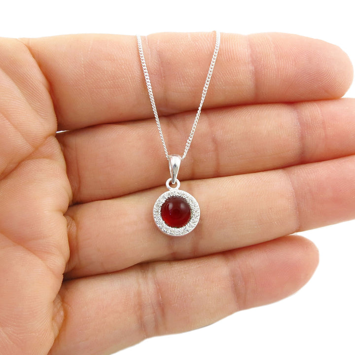 A sterling 925 silver pendant necklace with a round red carnelian agate cabochon gemstone surrounded by a halo of small clear stones, held in the palm of a woman's hand against a white background.
