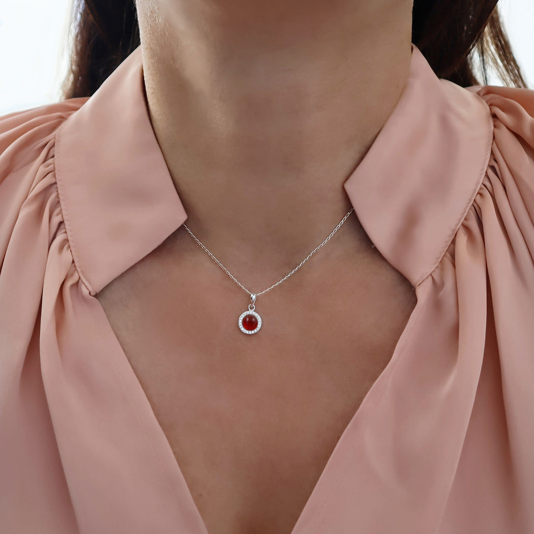 A close-up of a female model wearing a delicate 925 silver chain necklace with a circular pendant featuring a red carnelian agate gemstone and a halo of small clear stones, styled with a dusty pink blouse.