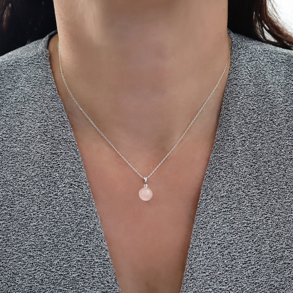 A close-up of a woman wearing a delicate silver chain necklace with a small circular pendant featuring a pink rose quartz gemstone centre styled with a gray textured top.