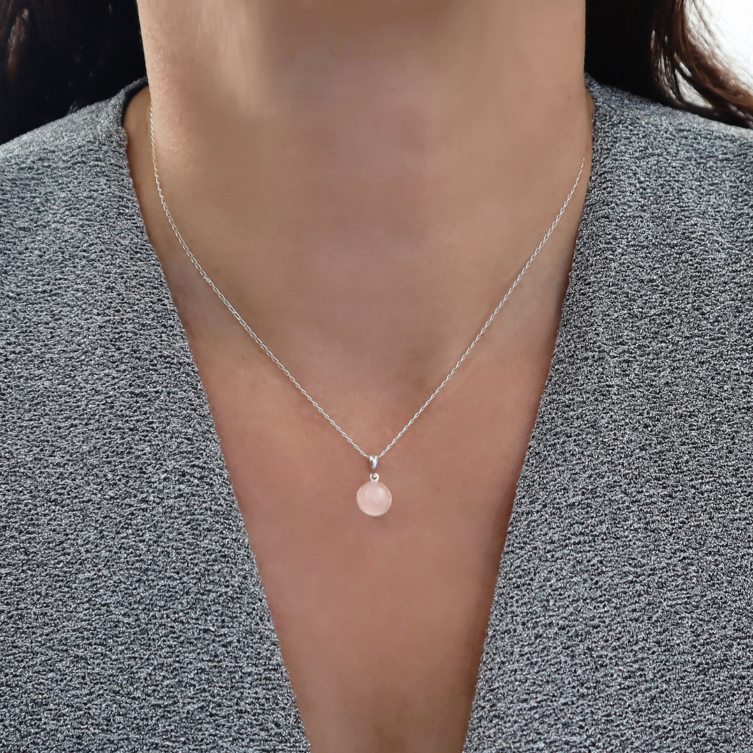 A close-up of a woman wearing a delicate silver chain necklace with a small circular pendant featuring a pink rose quartz gemstone centre styled with a gray textured top.