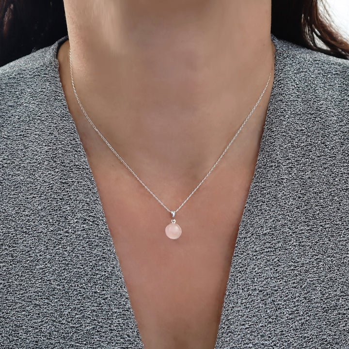 A close-up of a woman wearing a delicate silver chain necklace with a small circular pendant featuring a pink rose quartz gemstone centre styled with a gray textured top.