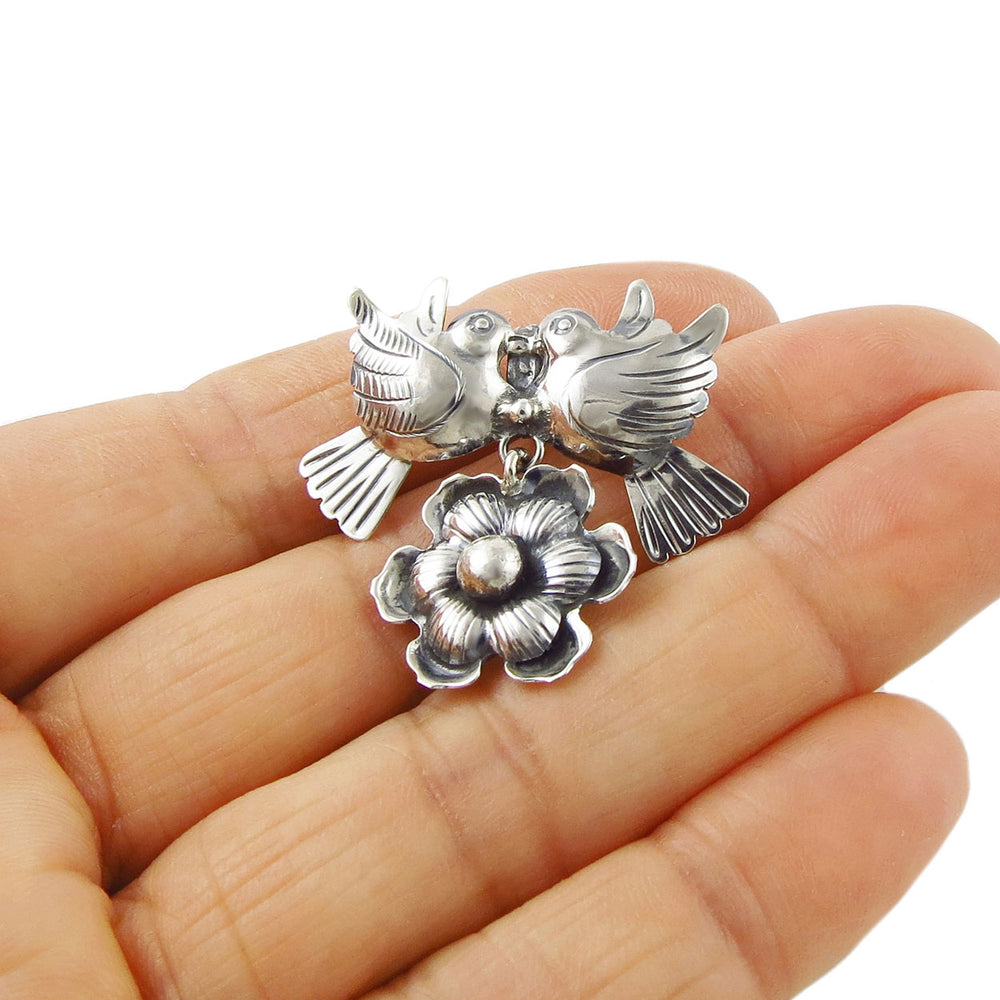 A sterling silver pendant featuring two lovebirds and a flower, held between thumb and forefinger.