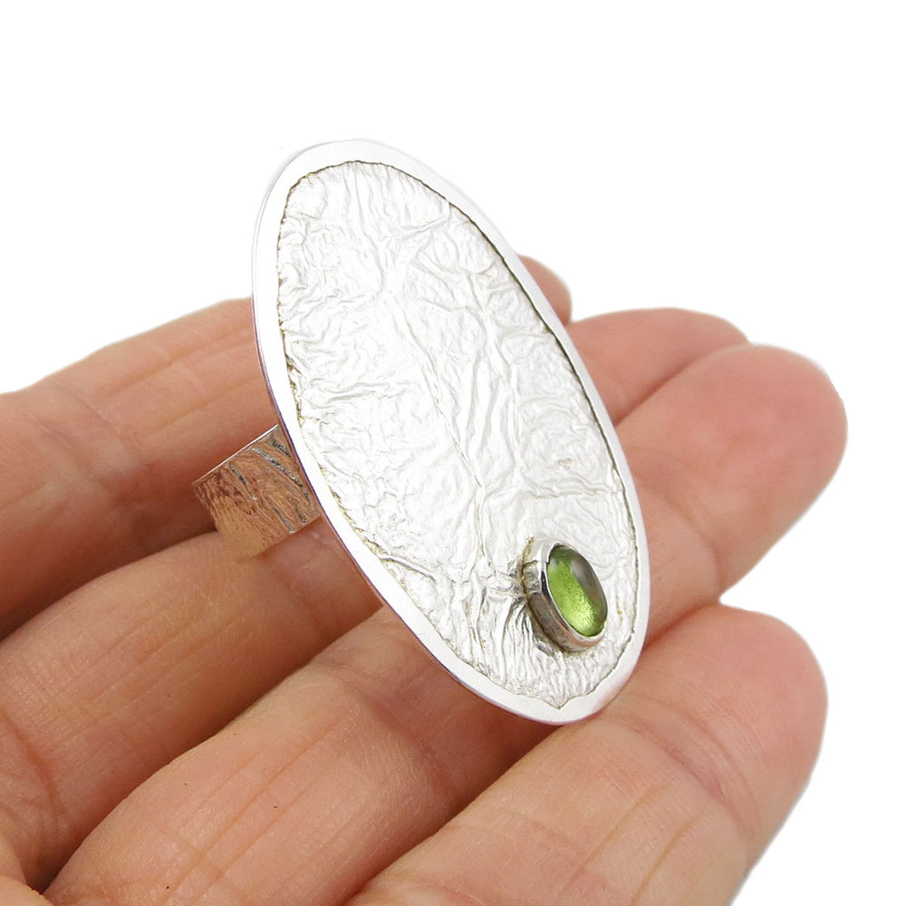 A large oval cocktail ring in crushed reticulated sterling silver with a peridot gemstone showcased in a woman's hand.  