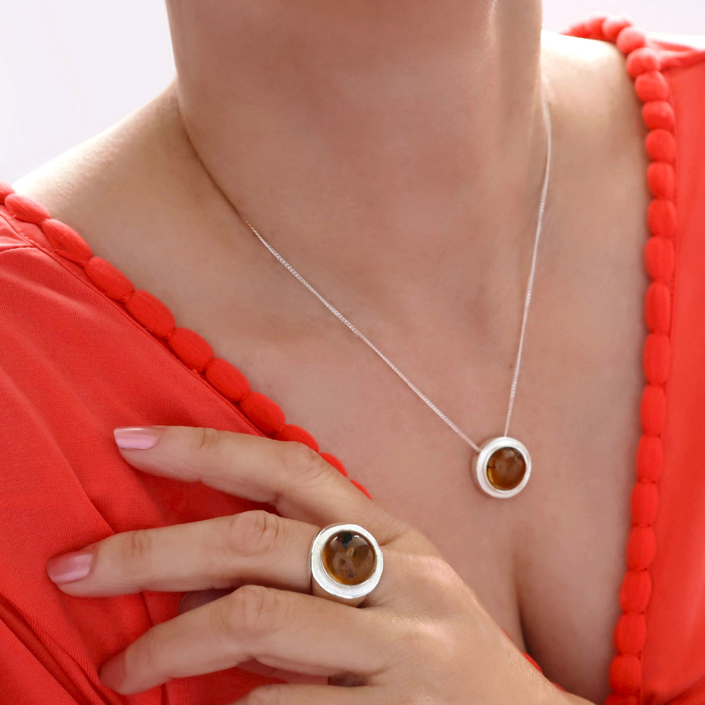 A woman wearing an orange dress showcases a silver ring with a honey-coloured amber gemstone on her finger and a matching necklace around her neck.