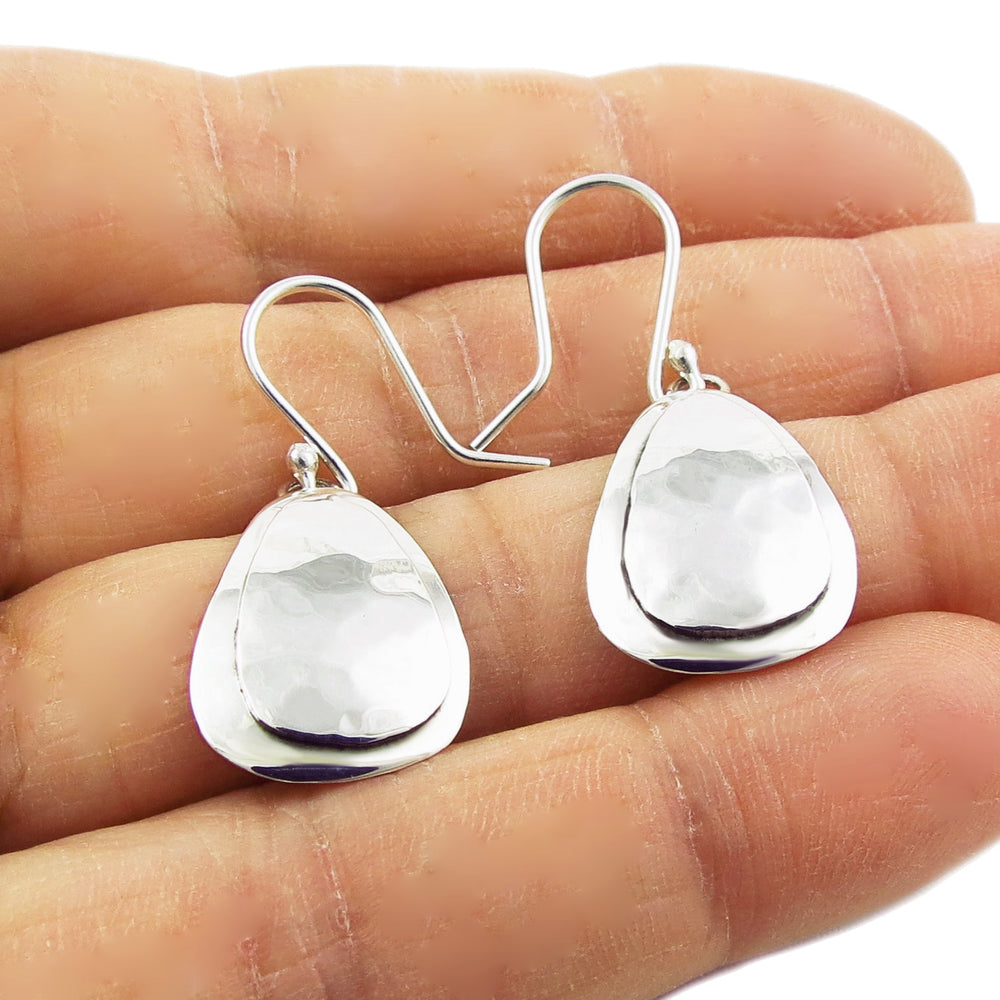 A pair of sterling silver teardrop earrings with a polished and hammered texture, displayed on a hand.
