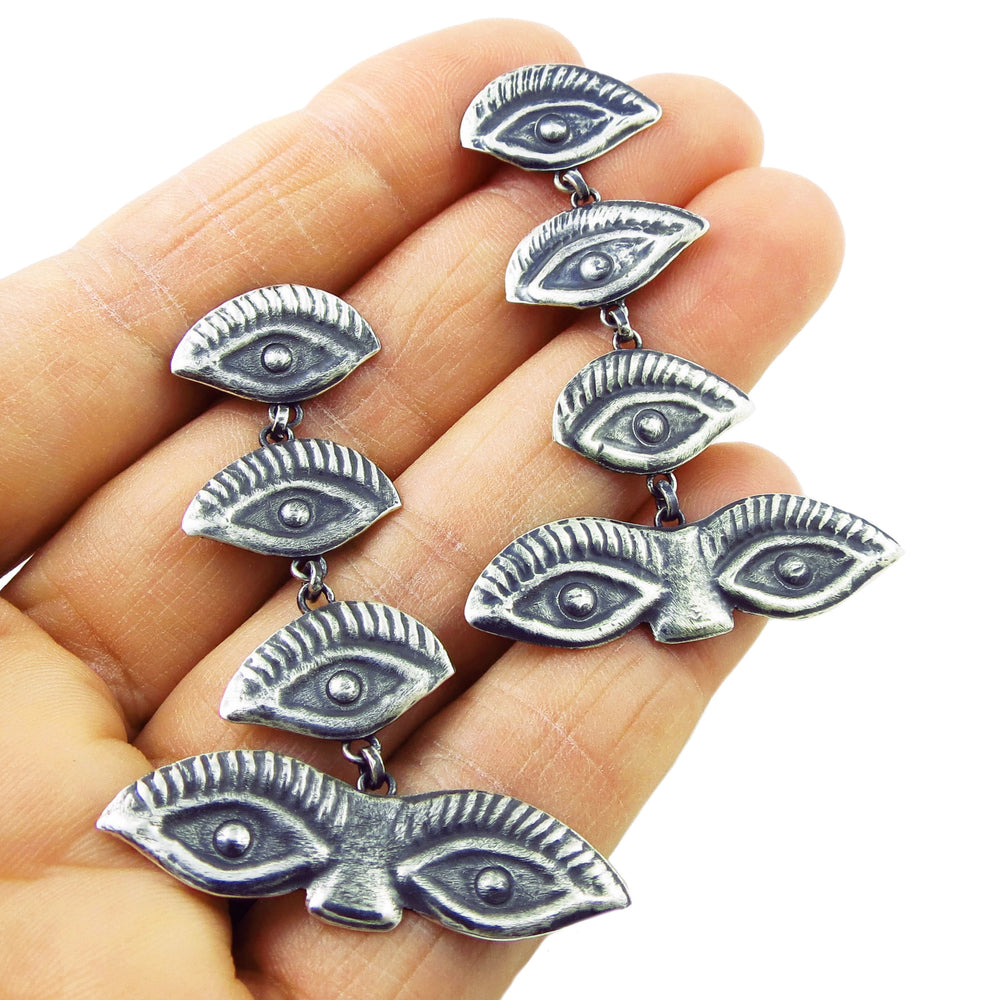 A pair of 925 sterling silver long drop earrings with a series of eye-shaped designs, held in a person's hand.