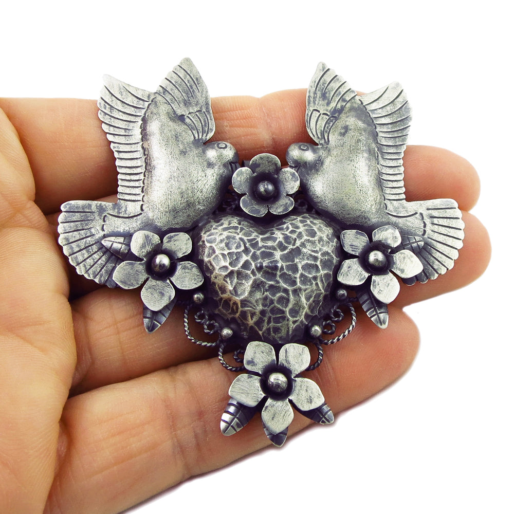 A large ornate Maria Belen Nilson designer 925 sterling silver Mexican lovebirds, flowers and heart pendant held in a person's hand
