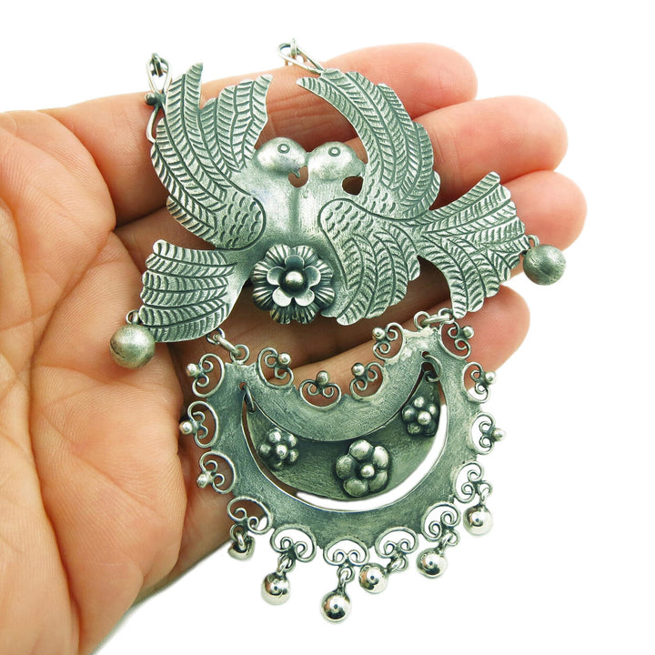 AA large Taxco 925 sterling silver bird and flower statement necklace with a blackened antiqued finish held in a woman's hand on a white background. 