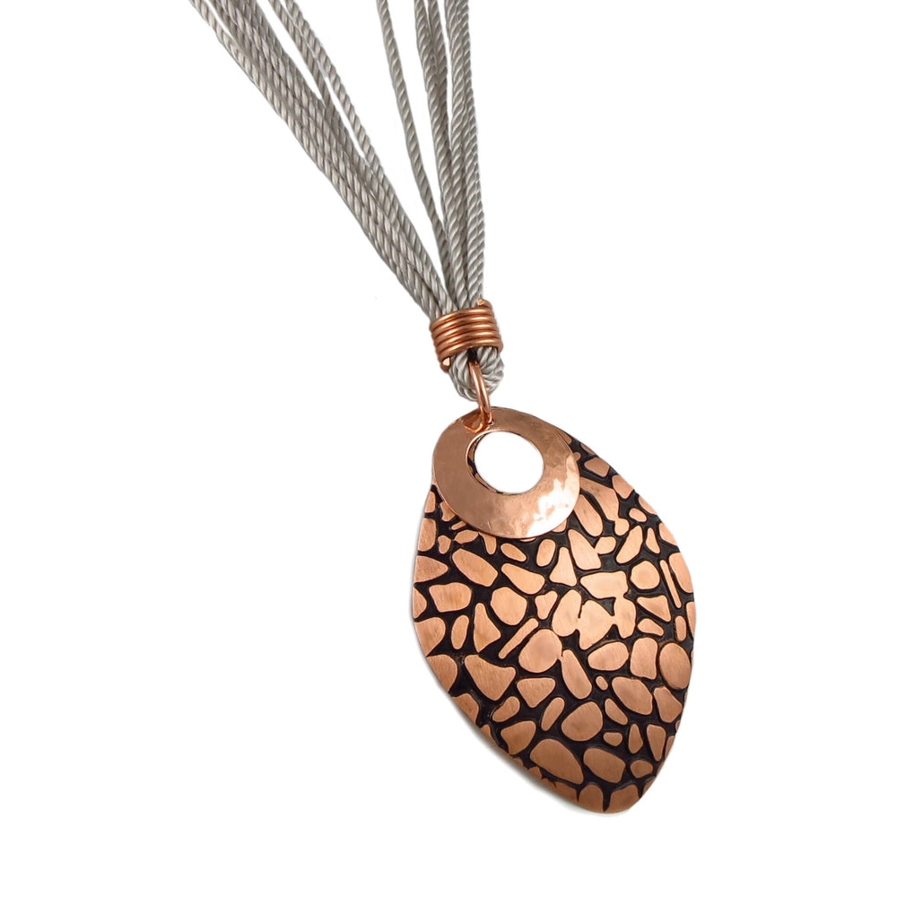 A large animal print copper pendant hanging on a grey multi rope cord necklace against a white background. 