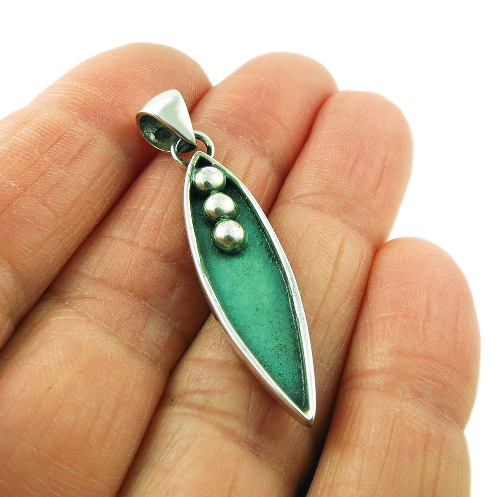 A sterling silver and copper pendant necklace designed as a pea pod, held between fingers.