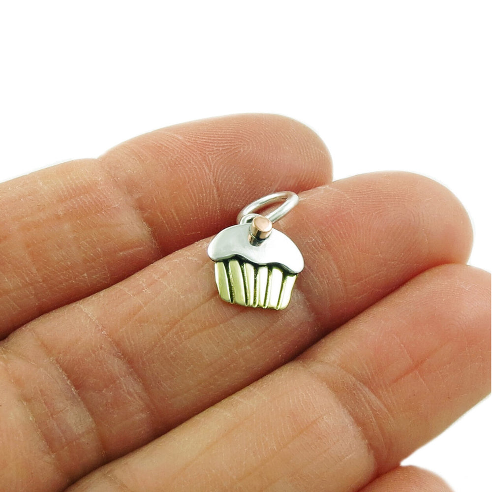 A small mixed metal cupcake pendant charm with a copper cherry on top, held between a woman's fingers.